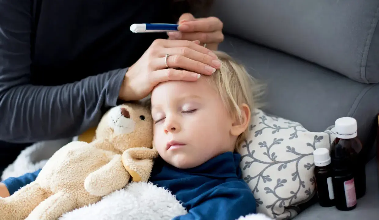 Bambino addormentato con febbre, la mamma controlla la temperatura. Vicino ci sono flaconi di antibiotico.
