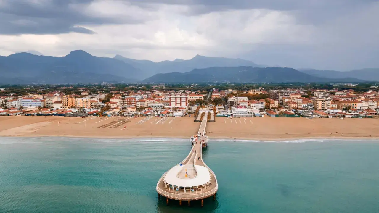 Un molo si estende su un mare azzurro, con una spiaggia dorata e una città costiera sullo sfondo. La Toscana offre un mare bello e panorami indimenticabili.