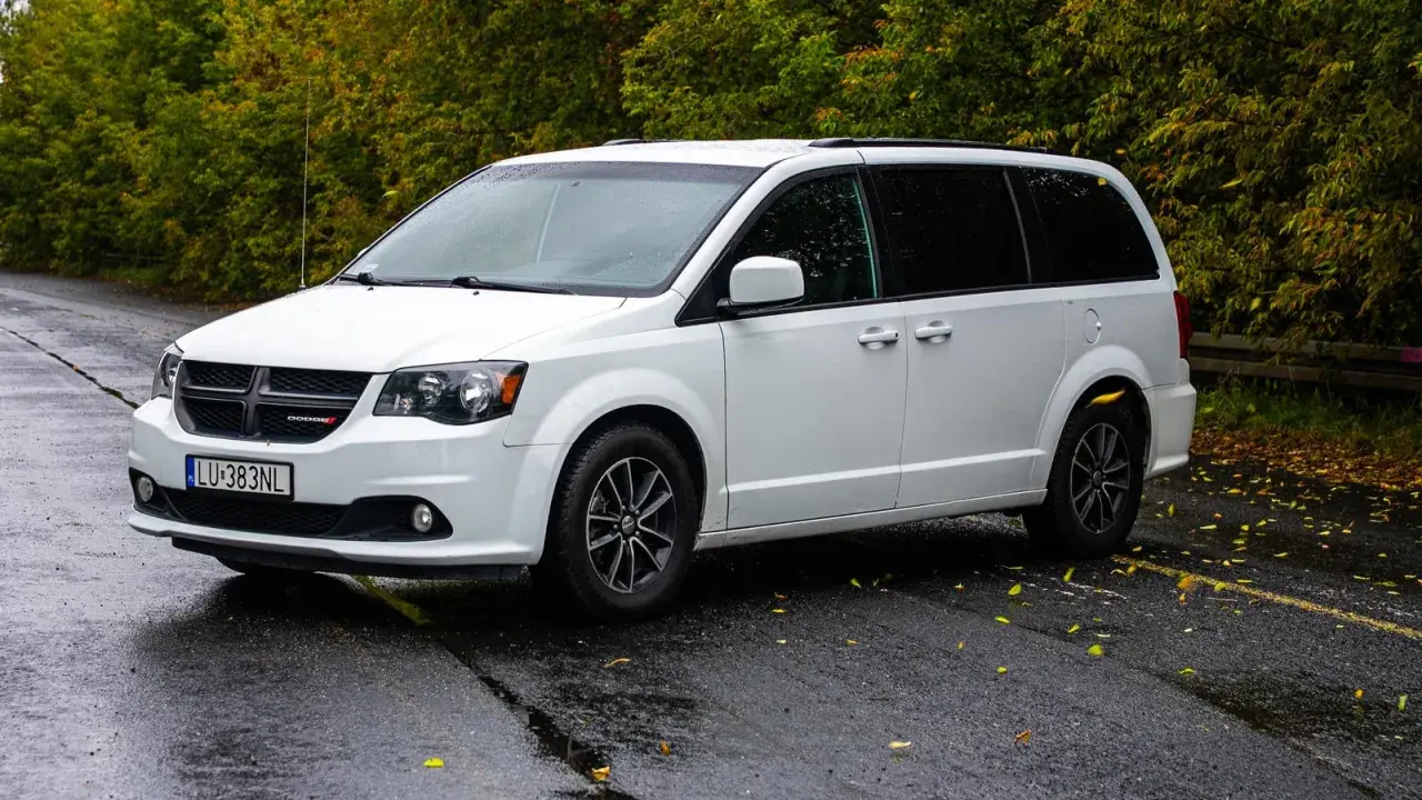 Biały Dodge Grand Caravan na mokrej drodze, otoczony jesienną zielenią.