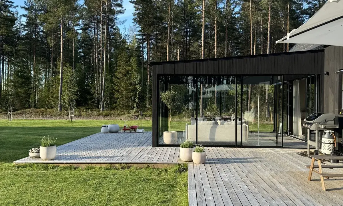 Enkel uteplats med trätrall och glaspartier som vetter mot en grönskande skog. Perfekt för avkoppling.