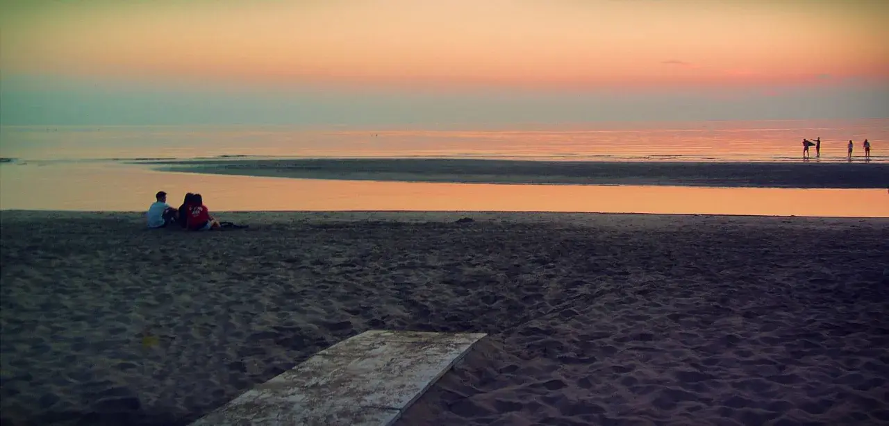 Alba a Rimini: una coppia seduta sulla sabbia guarda il mare calmo e il cielo colorato.