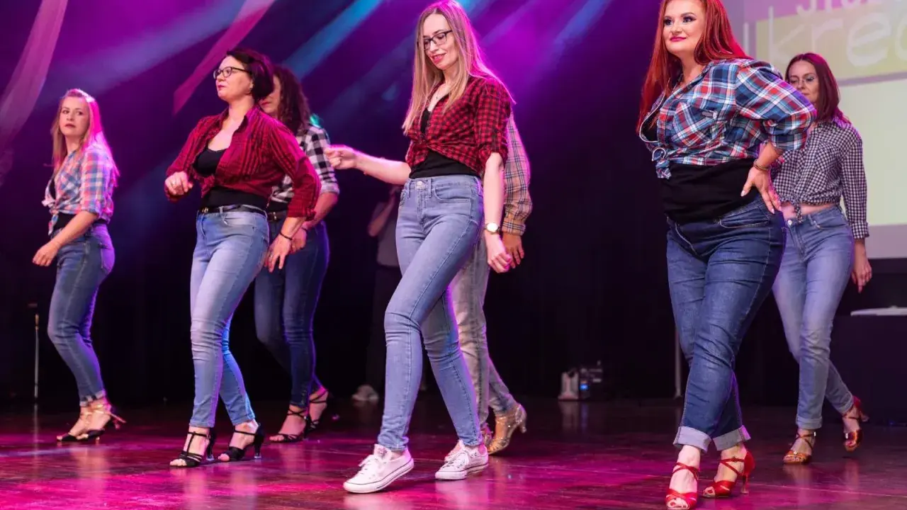 Grupa kobiet w kratę i jeansach tańczy na scenie. Występ latino solo dla początkujących.