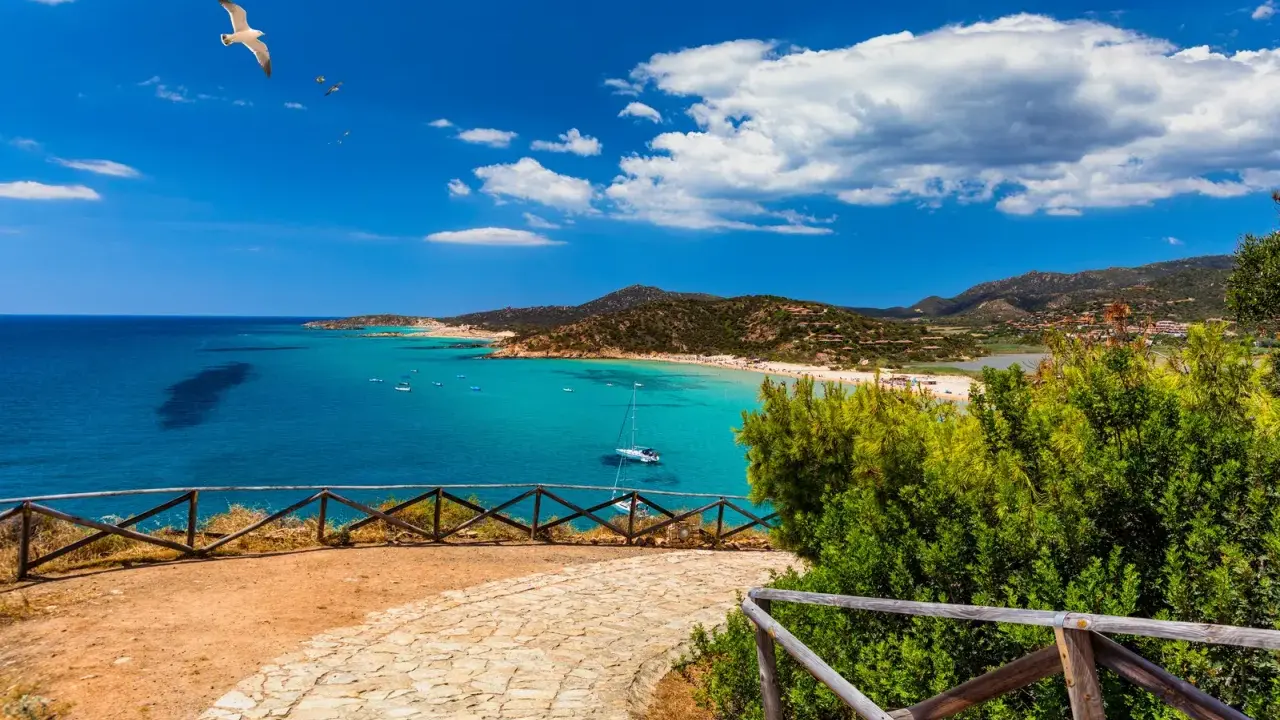 Vista panoramica di una baia con acque turchesi, spiagge affollate e montagne verdi. Un gabbiano vola nel cielo azzurro sopra Quartu e dintorni.