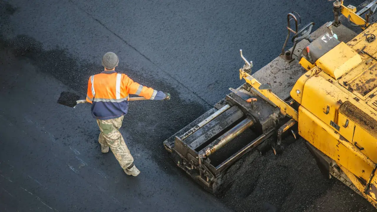 Arbeiter schaufelt heißes Bitumen auf eine Straße, während ein Asphaltfertiger das Material verteilt.