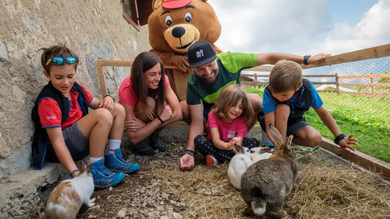 Bambini e adulti interagiscono con conigli in un recinto, parte di un'esperienza in fattorie didattiche. Un personaggio con costume da orso sorride.