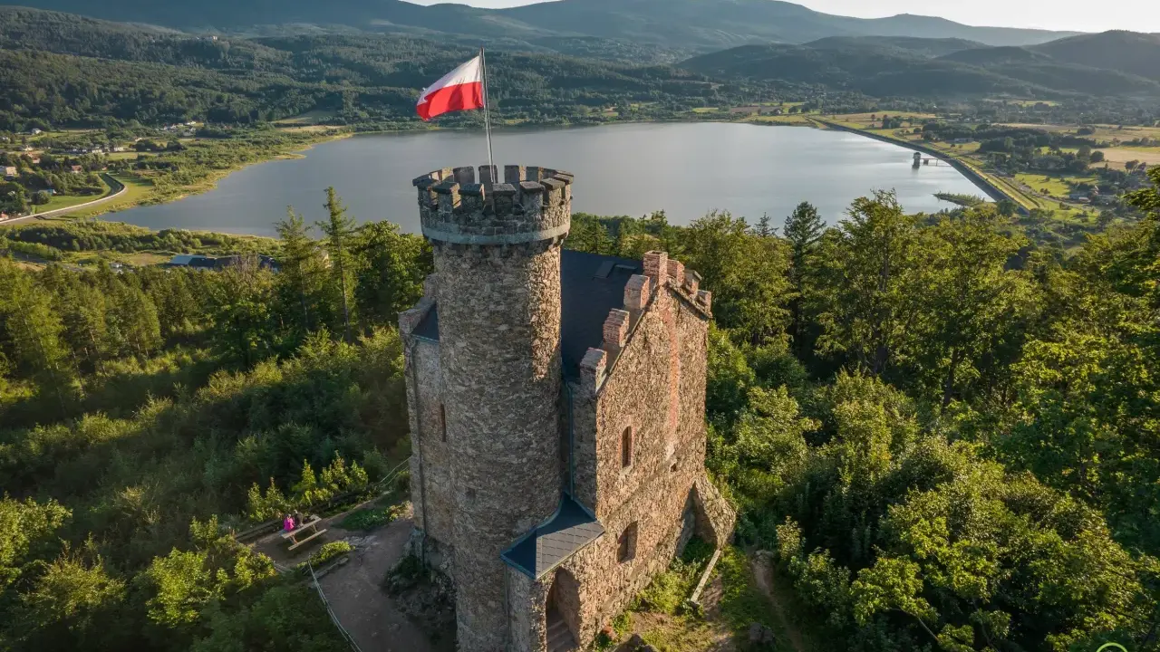 Kamienna wieża z polską flagą na tle jeziora i gór. Wspaniałe **cieplice atrakcje** dla miłośników historii i przyrody.