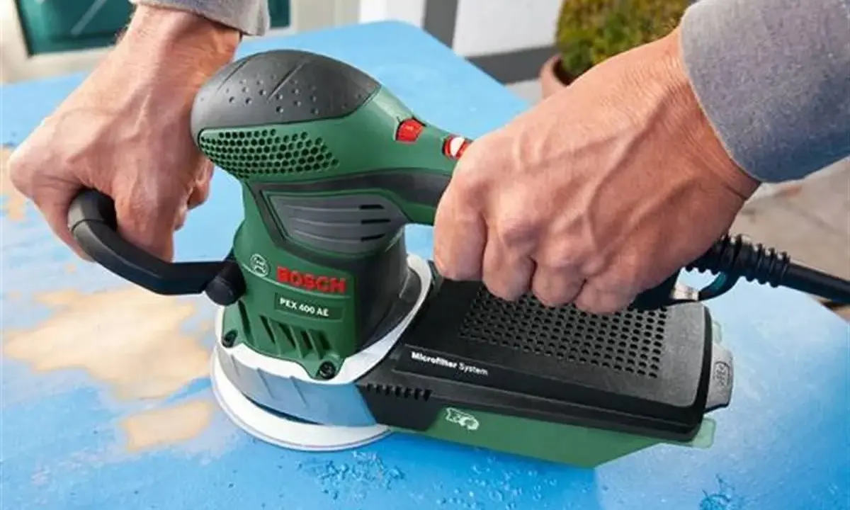 Dłonie trzymają zieloną szlifierkę Bosch PEX 400 AE. To szlifierka oscylacyjna a mimośrodowa, która usuwa starą farbę z niebieskiej powierzchni.