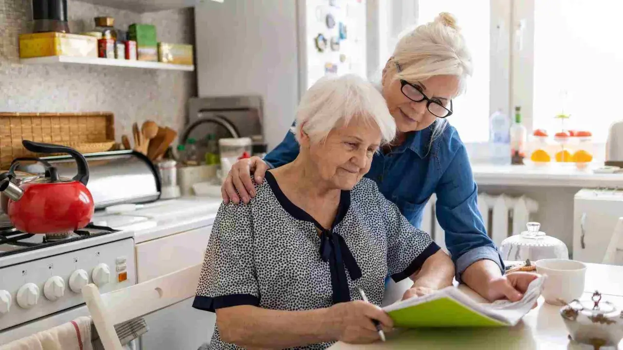 Dos mujeres mayores revisan documentos en la cocina. Una apoya a la otra, quizás tramitando la pensión de orfandad mayores de 30 años.