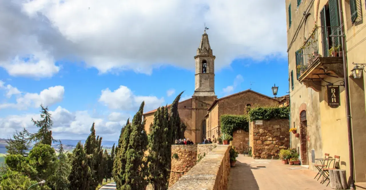 Un borgo nei comuni Val d'Orcia: un campanile svetta tra cipressi e case antiche, con un bar all'aperto.