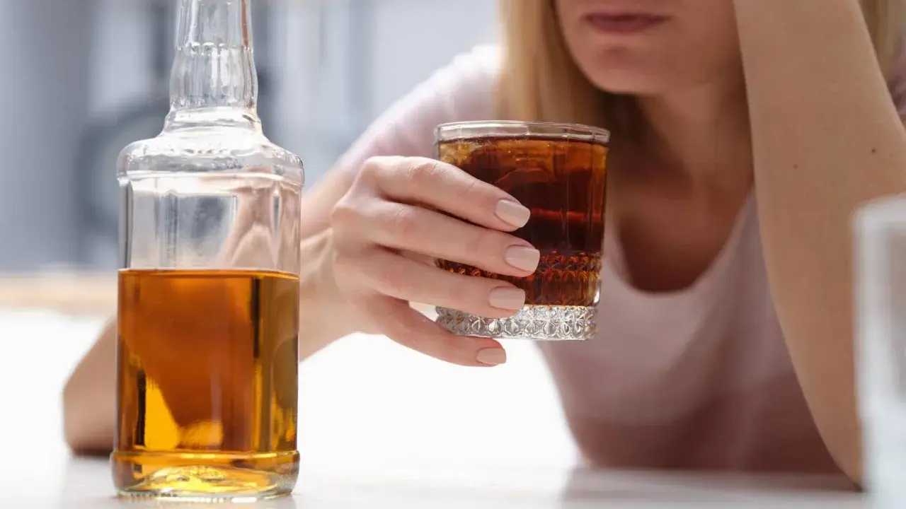 Kobieta z kieliszkiem drinka i butelką alkoholu. W tle widać jej twarz, która wyraża smutek. To może być wszywka alkoholowa.