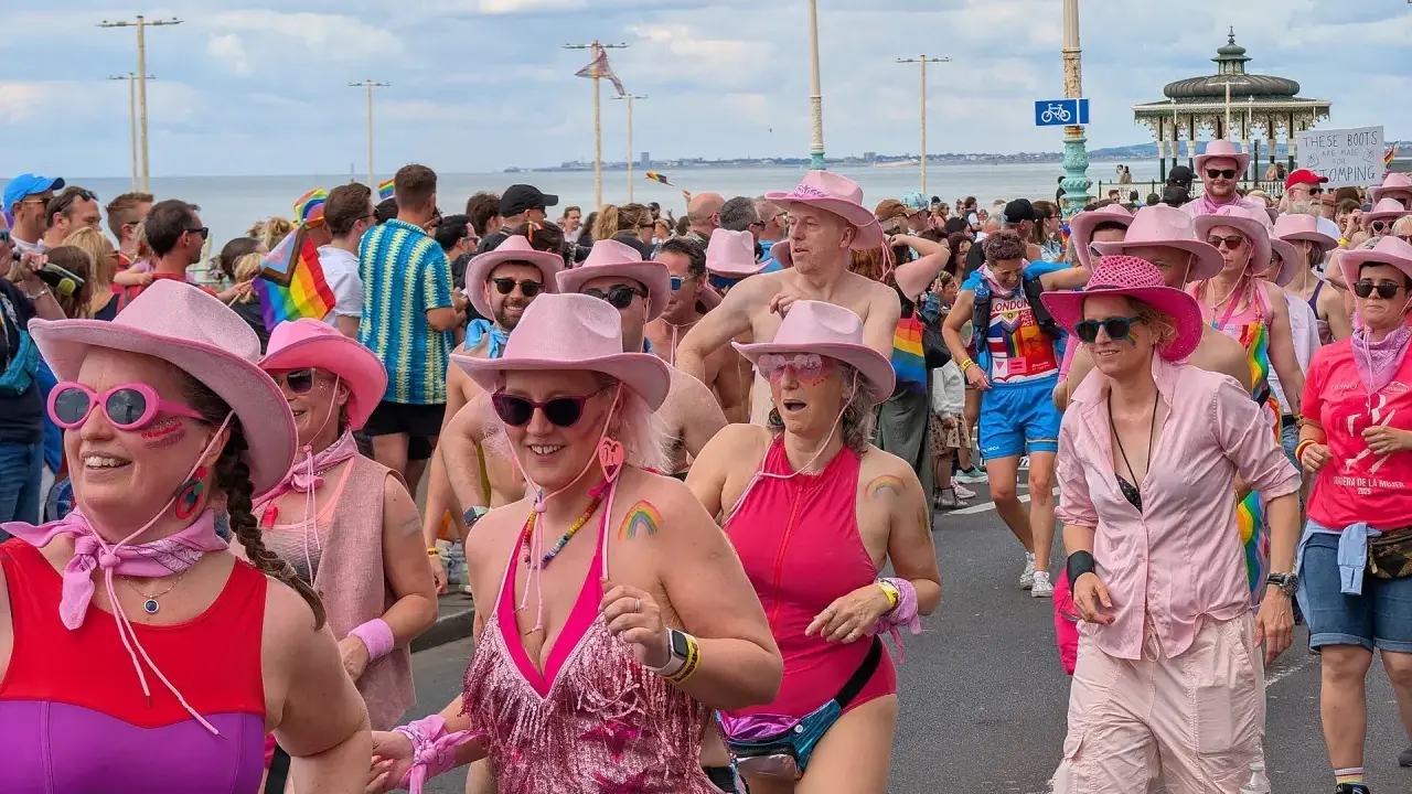A vibrant Brighton gay pride parade, with many participants wearing pink cowboy hats and rainbow-themed attire, celebrating joyfully by the sea.