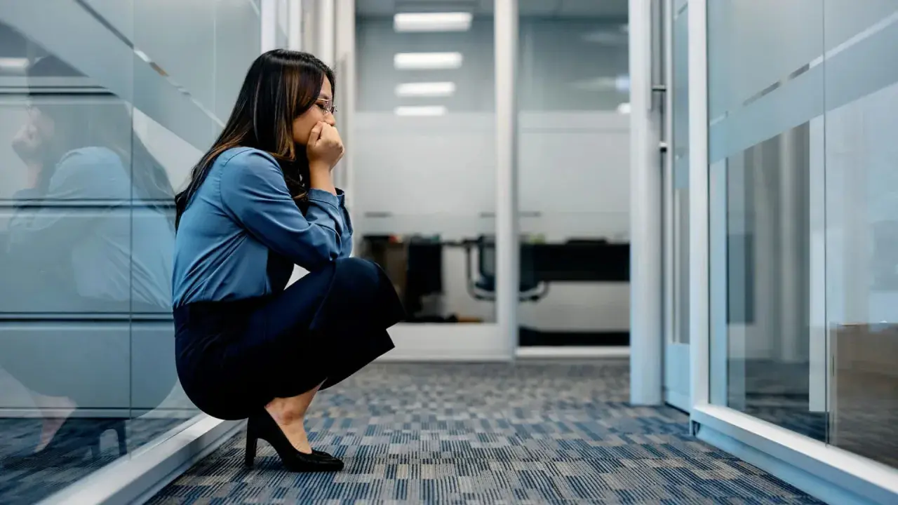 Frau kauert im Büroflur, Hände vor dem Mund, die Augen geschlossen. Sie scheint von **Angst vor der Arbeit** überwältigt zu sein.