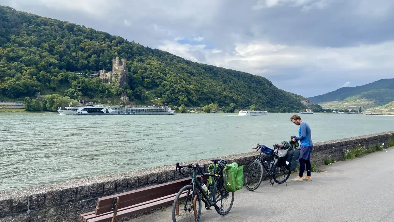 Mann packt Gepäck für eine Fahrradtour am Rhein. Im Hintergrund Burg und Fluss.