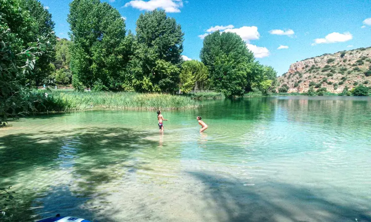 Dos niños juegan en las cristalinas aguas de las Lagunas de Ruidera, rodeados de vegetación y un paisaje montañoso.