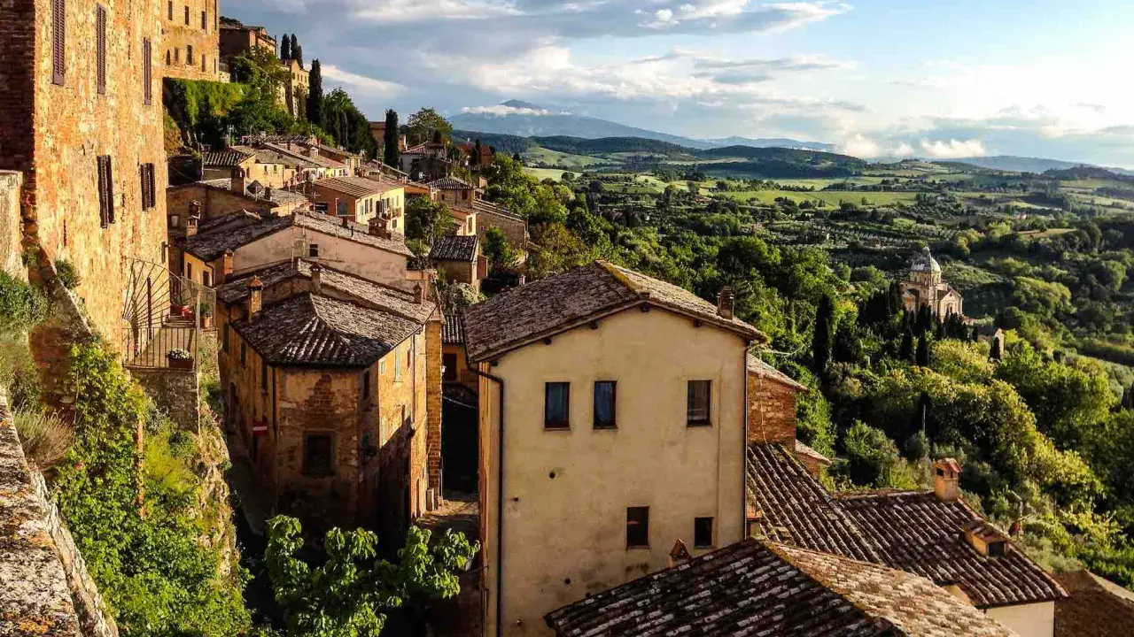 Panoramica di borghi toscana in treno, con case in pietra e tegole rosse su colline verdi sotto un cielo nuvoloso.