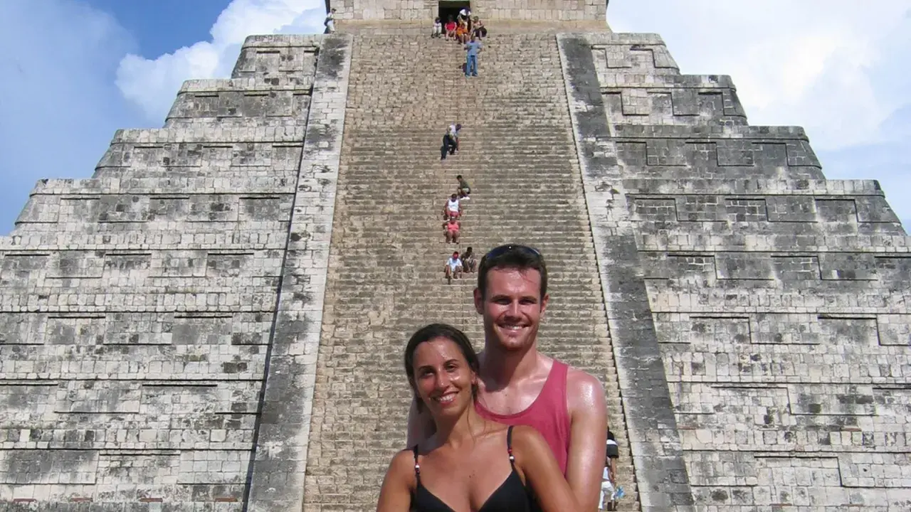 Coppia sorridente davanti alla piramide di Kukulkan, con turisti che salgono le scale del chichen itza interno.