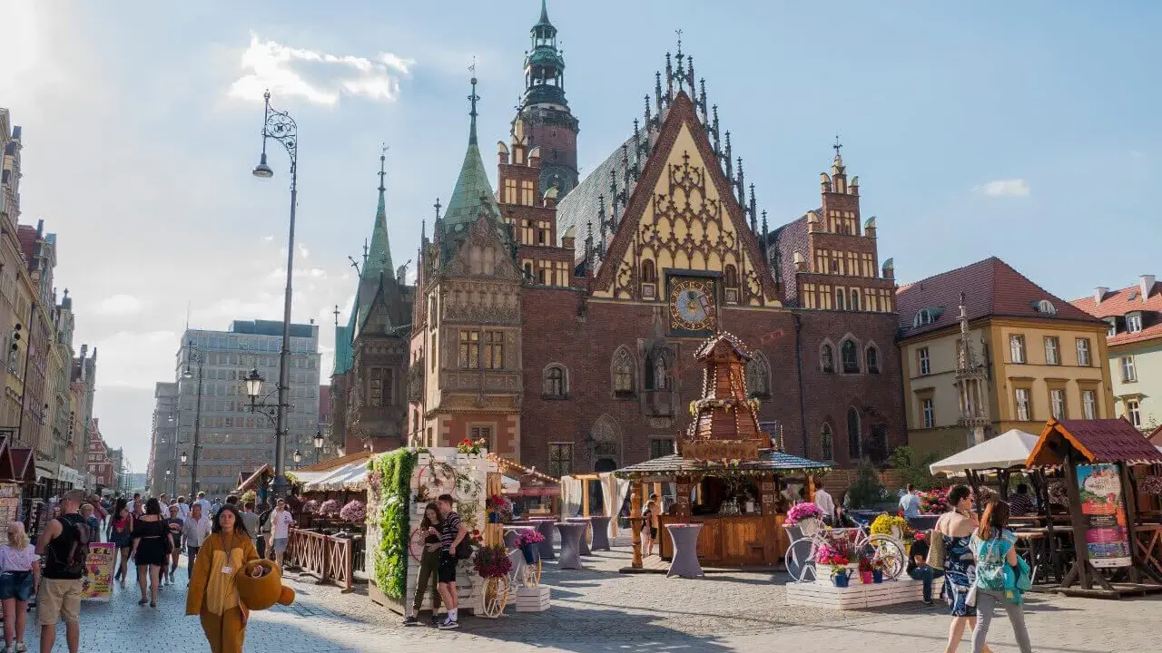 Tłum ludzi spaceruje po Rynku we Wrocławiu, podziwiając zabytkowy ratusz i letnie stoiska.