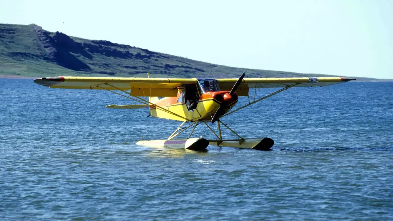 Gelbe Piper Super Cub mit Schwimmern auf ruhigem Wasser, im Hintergrund eine grüne Hügelkette.