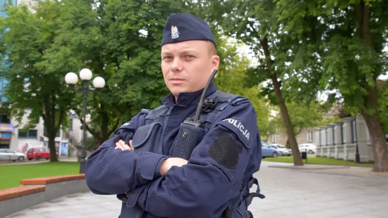 Policjant w granatowym mundurze, w sprawie umundurowania policjantów, stoi na tle zieleni.