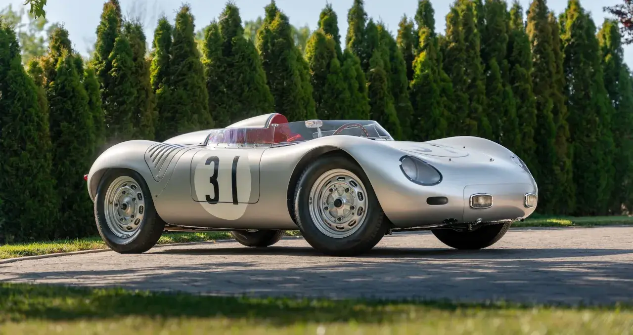 Silberner Porsche 718 RSK mit Startnummer 31, bereit für die Rennstrecke, vor einer Reihe von grünen Bäumen.