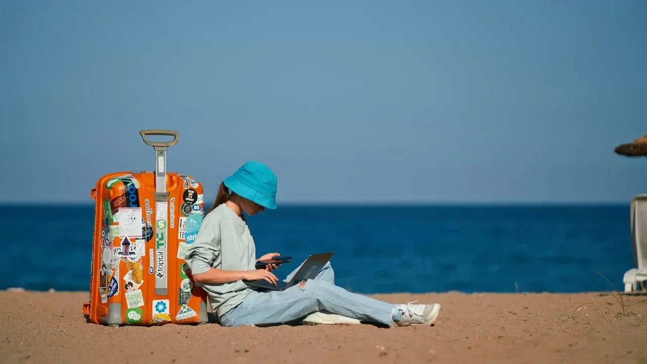Dziewczyna w niebieskim kapeluszu siedzi na plaży z walizką, laptopem i telefonem, planując, jak tanio podróżować.
