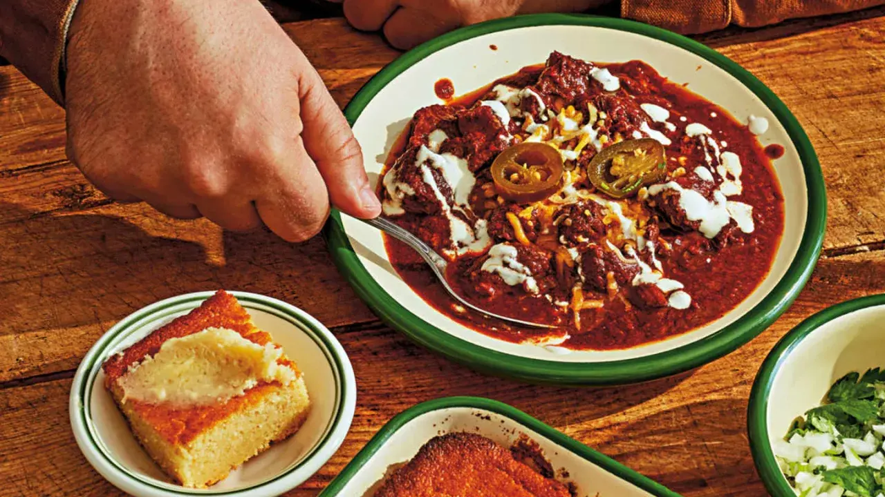 En hand tar en sked ur en skål med mustig texas chili, toppad med jalapeños och gräddfil. Bredvid ligger gyllene majsbröd.