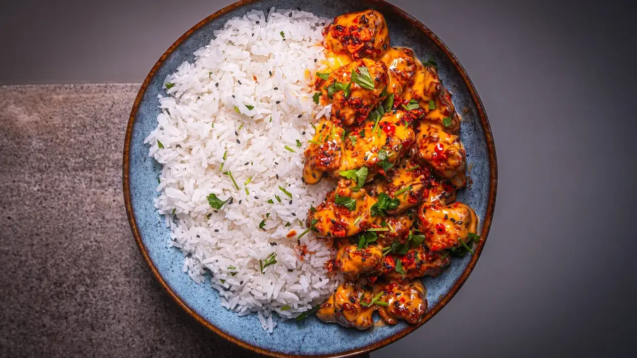Schüssel mit weißem Reis und würzigem Bang Bang Chicken, garniert mit Koriander und Sesam.