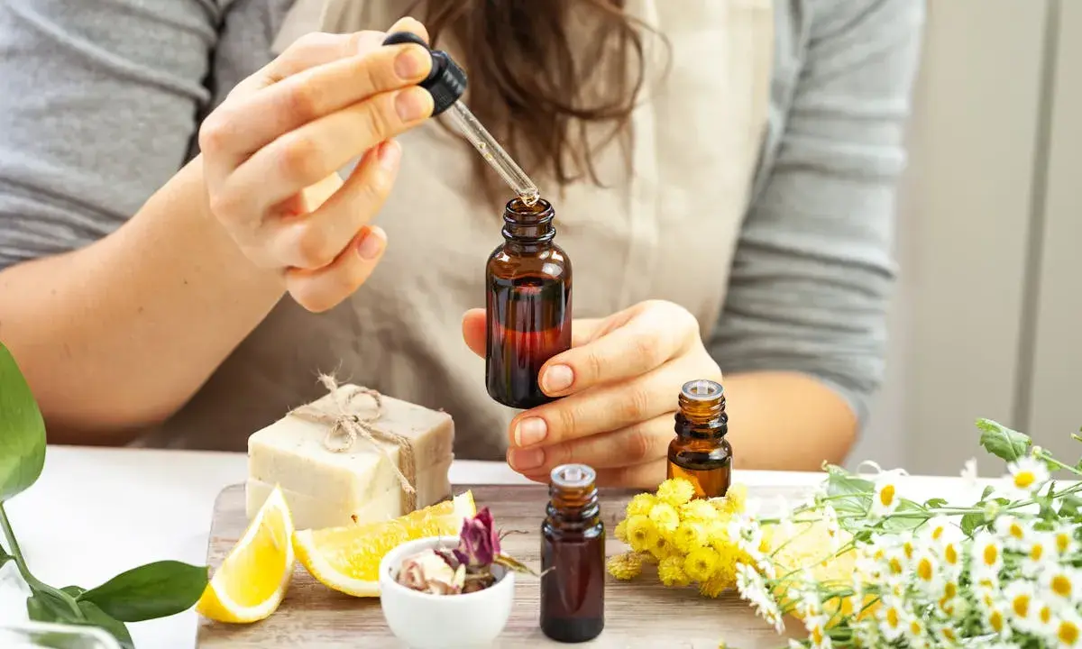 Une personne prépare un parfum huile essentielle, ajoutant une goutte d'ambre dans un flacon. Des fleurs, des citrons et du savon sont disposés autour.