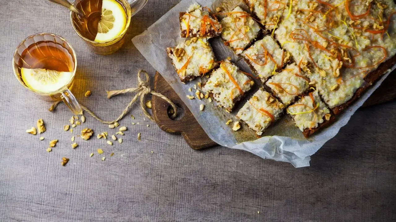 En skiva vegansk morotskaka med glasyr och nötter, serverad med två koppar te med citron.