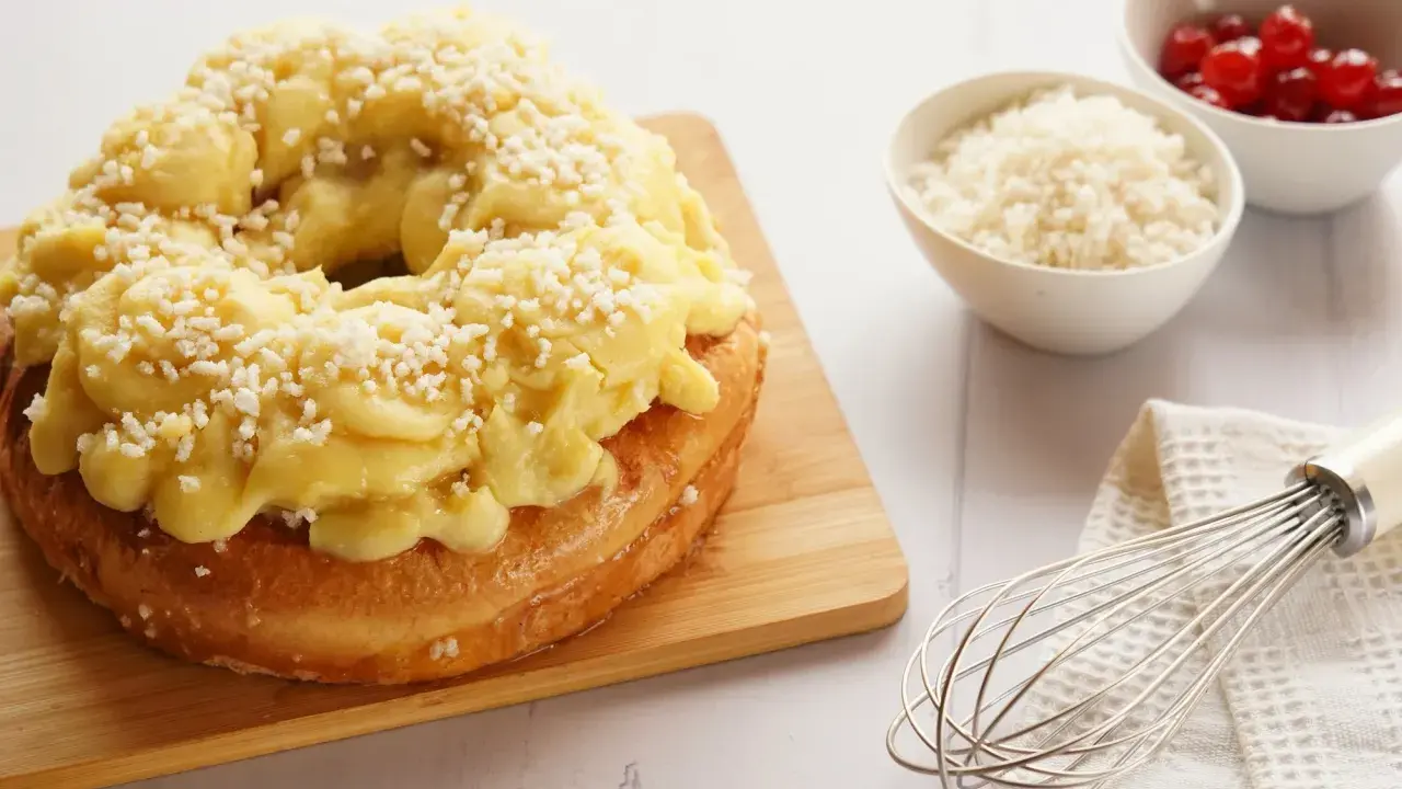 Rosca de Pascua decorada con crema pastelera y azúcar granulada, lista para disfrutar. A su lado, cuencos con cerezas y ralladura, y un batidor.