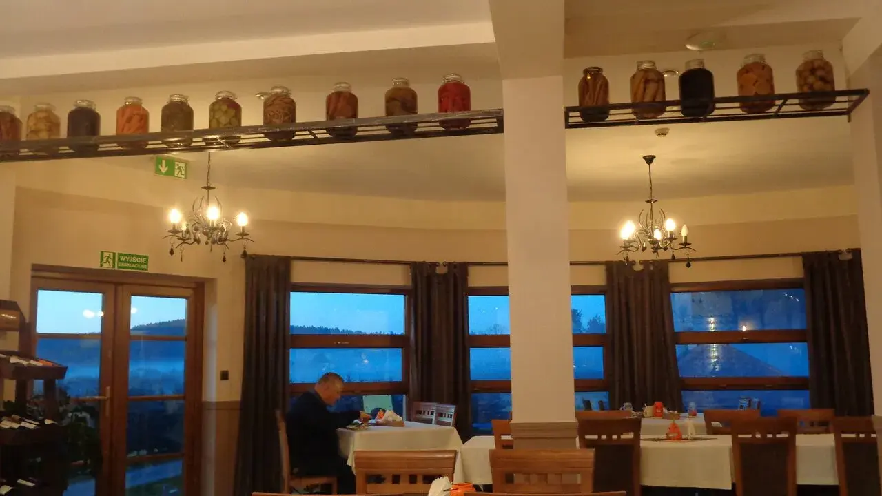 Wnętrze restauracji z oknami wychodzącymi na góry, stoły nakryte obrusami, a na półce nad nimi słoiki z przetworami.