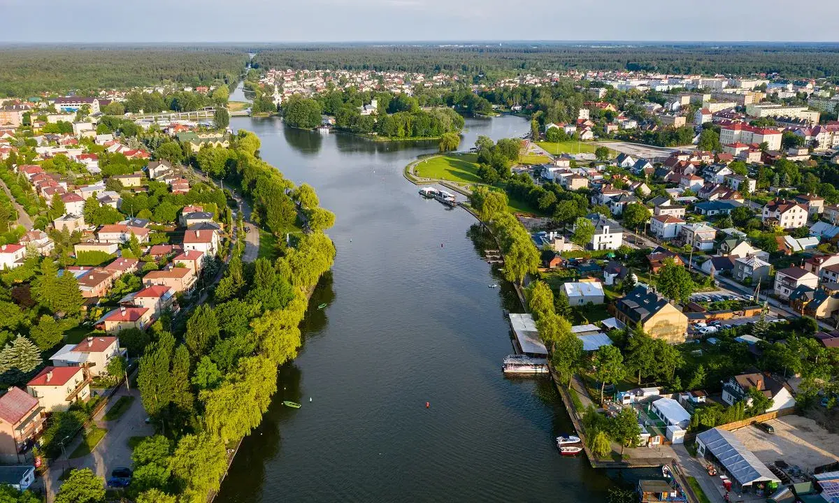 Malowniczy widok na Augustów i jego atrakcje: rzeka, zielone brzegi, domy i las w tle.