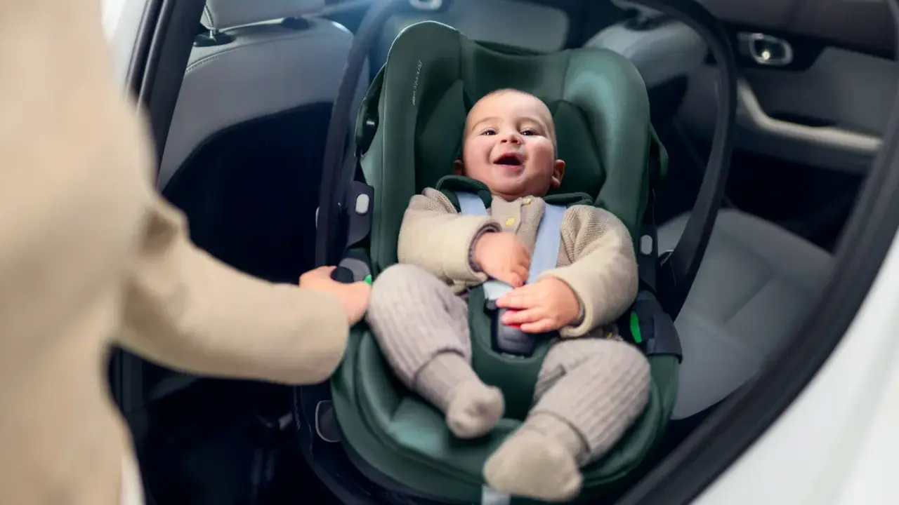 Uśmiechnięte dziecko w foteliku samochodowym. Ręka rodzica pokazuje, jak wypiąć fotelik z bazy isofix.