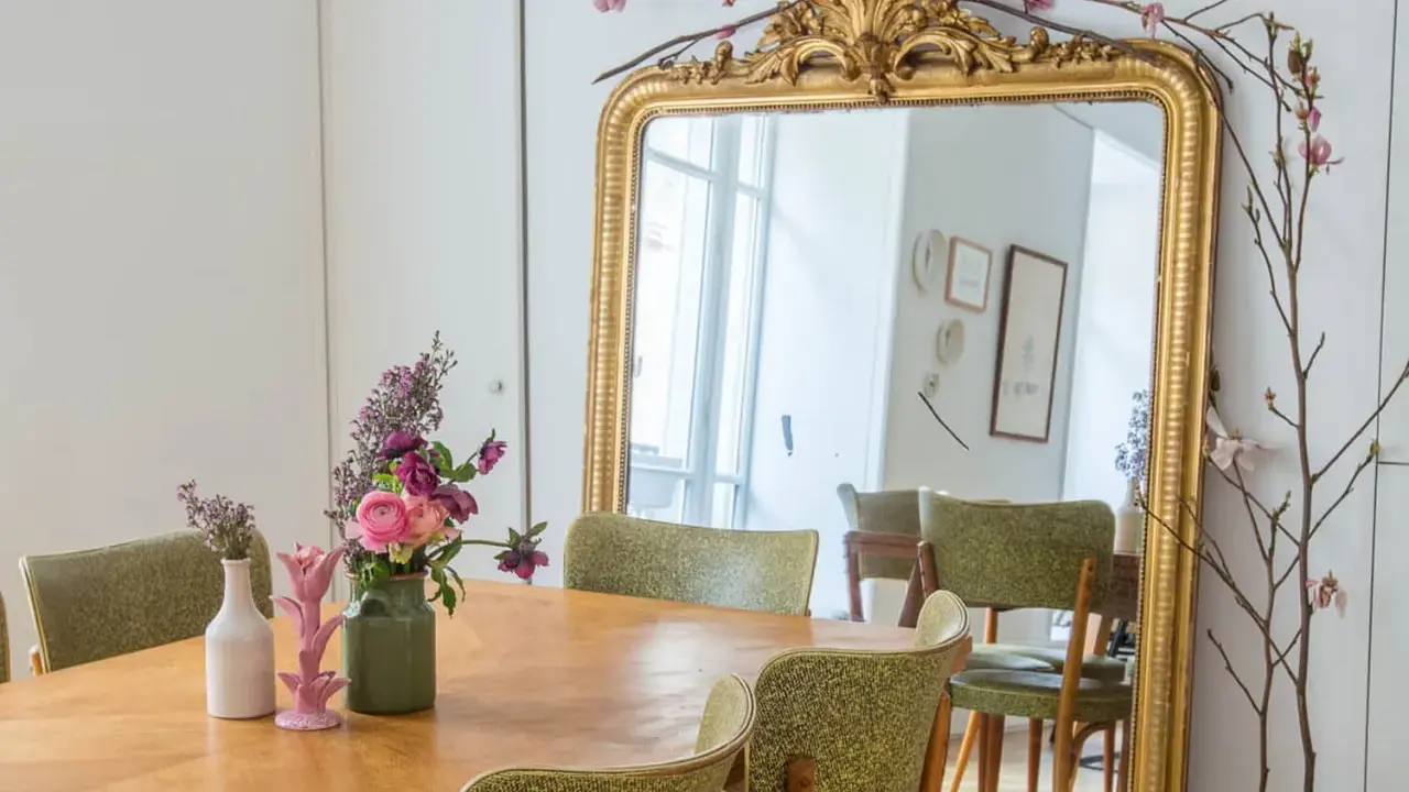 Un grand miroir ancien doré, orné de branches fleuries, reflète une salle à manger avec des chaises vertes. Quelle couleur peindre un miroir ancien ? L'or reste un classique intemporel.