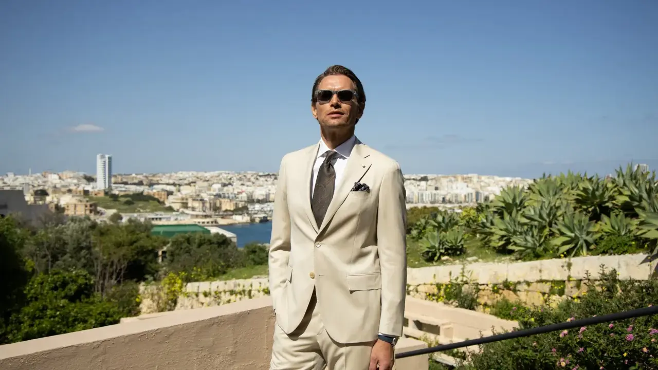 A man in a light tan suit, tie, and sunglasses offers inspiration for what to wear to a communion as a guest male.
