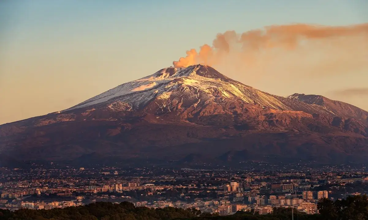Etna fumante, con la cima innevata che brilla al tramonto, domina una città siciliana.