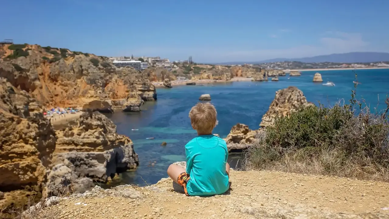 Kleinkind blickt auf die Küste Portugals. Felsen, türkisfarbenes Meer und eine Bucht mit Strand.