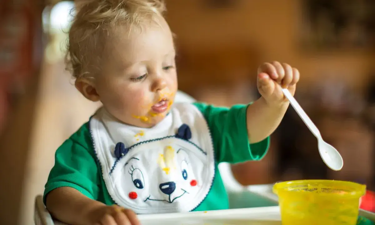 Małe dziecko z marchewkowym puree na buzi i śliniaczku próbuje samodzielnie jeść. Oto jak sprać marchewkę z ubranka!