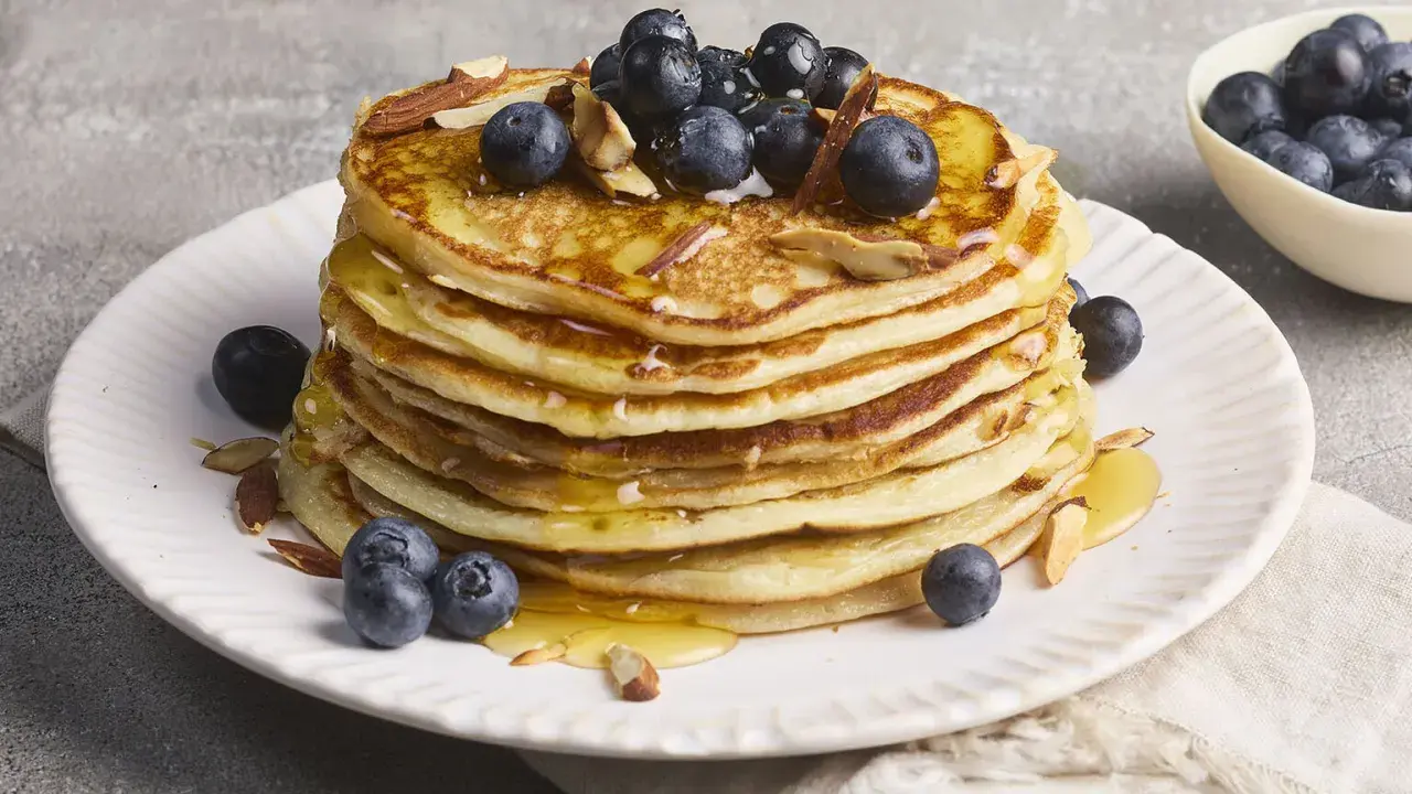 Stapel fluffiger High Protein Pancakes mit Blaubeeren und Mandelsplittern, beträufelt mit Honig.