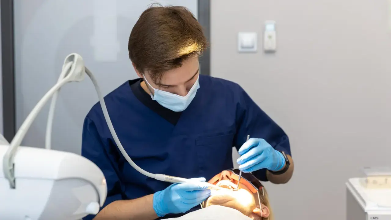 Dentysta w niebieskim fartuchu i masce wykonuje zabieg. Pacjentka ma założone okulary ochronne. Nowoczesne znieczulenie komputerowe zapewnia komfort.