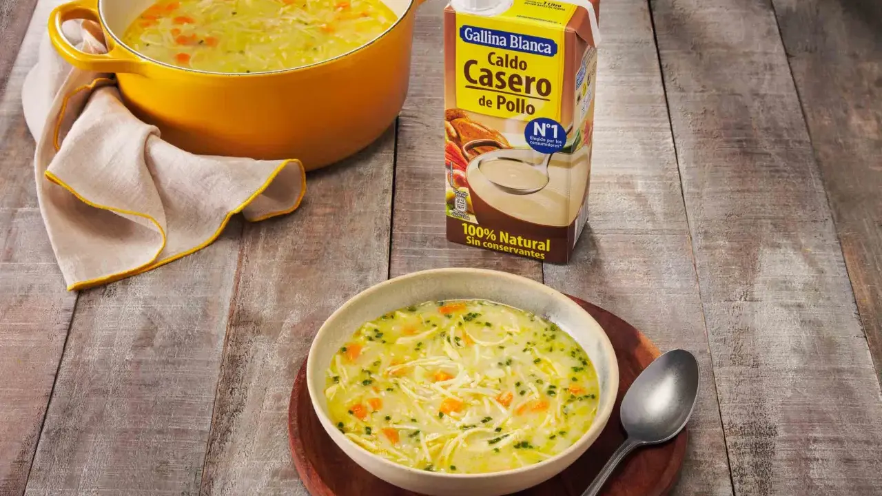 Un tazón humeante de sopa de pollo casera, con fideos finos y trozos de zanahoria, junto a una olla amarilla y un cartón de caldo de pollo.