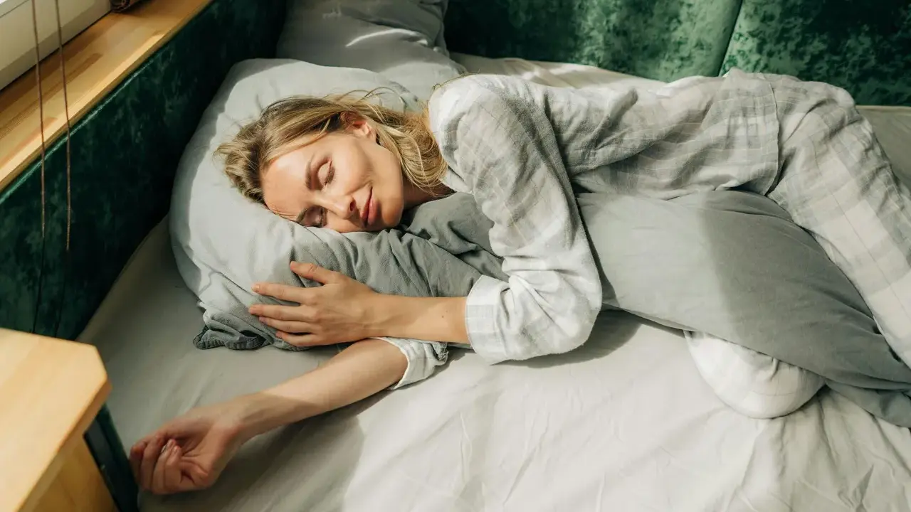 En kvinna sover lugnt i sängen, omfamnande en kudde. Solstrålar faller in genom fönstret och skapar en känsla av guidad avslappning.