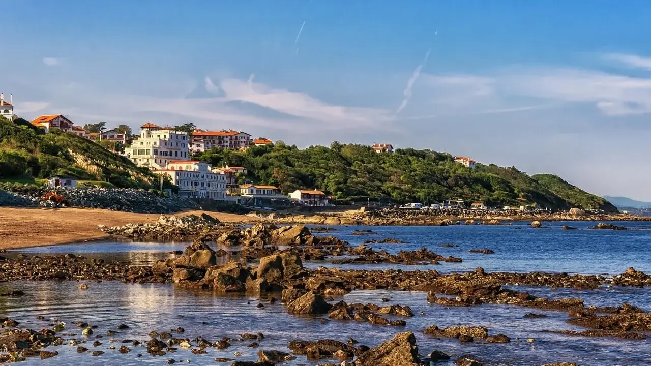 Vue côtière avec des maisons blanches sur une colline verdoyante, une plage de sable et des rochers dans l'eau. Le cadastre zone inondable est visible.