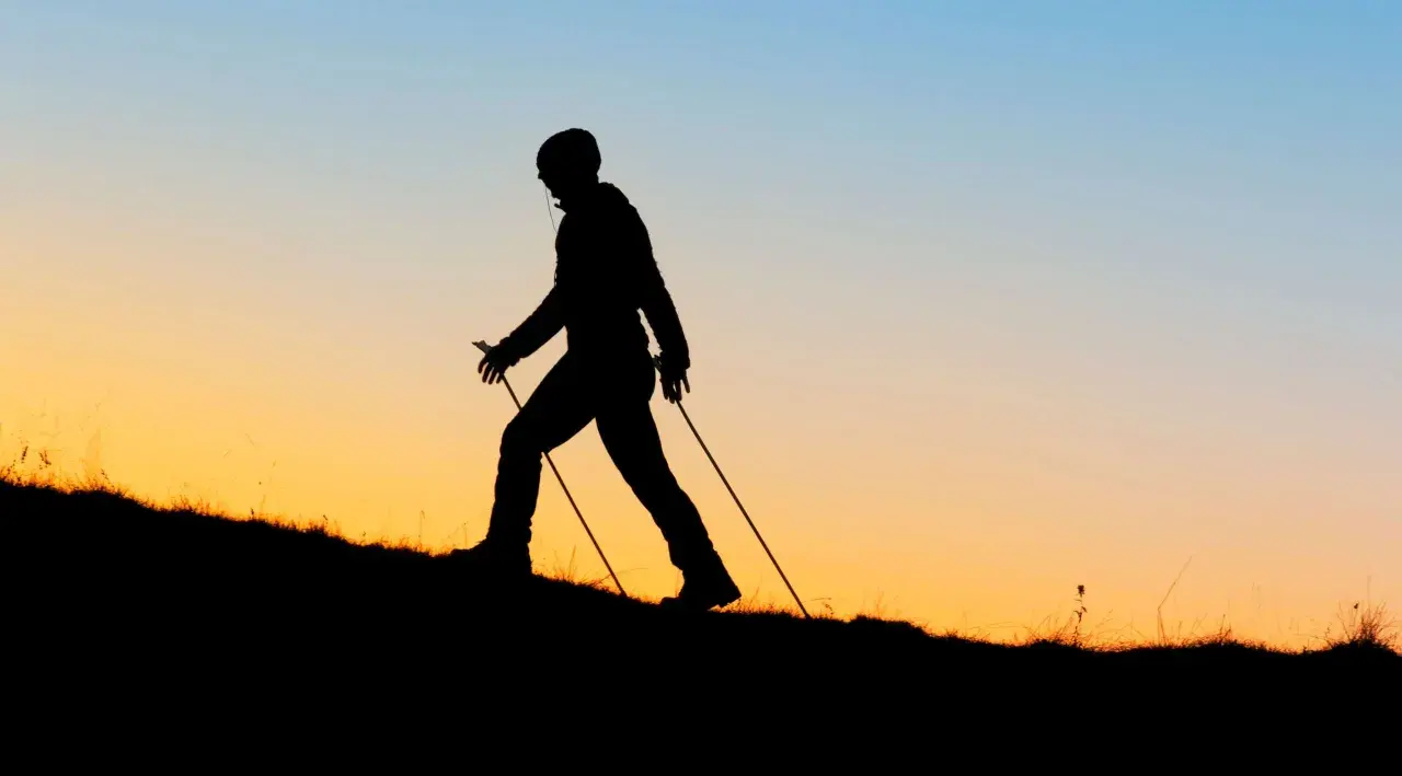 Sylwetka osoby uprawiającej nordic walking na tle zachodzącego słońca. Czy wolisz nordic walking rano czy wieczorem?