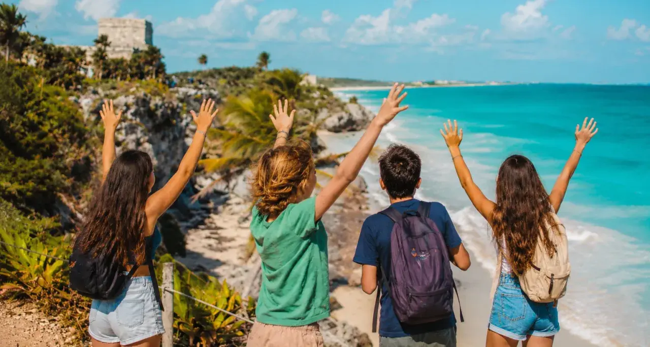Cuatro jóvenes con mochilas saludan al mar turquesa y las ruinas mayas. ¡Una aventura inolvidable en la Riviera Maya con niños!
