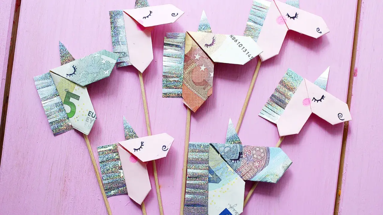Kreatives Geld falten: Mehrere Einhorn-Figuren aus Geldscheinen und Glitzerpapier auf Holzstäbchen vor rosafarbenem Hintergrund.
