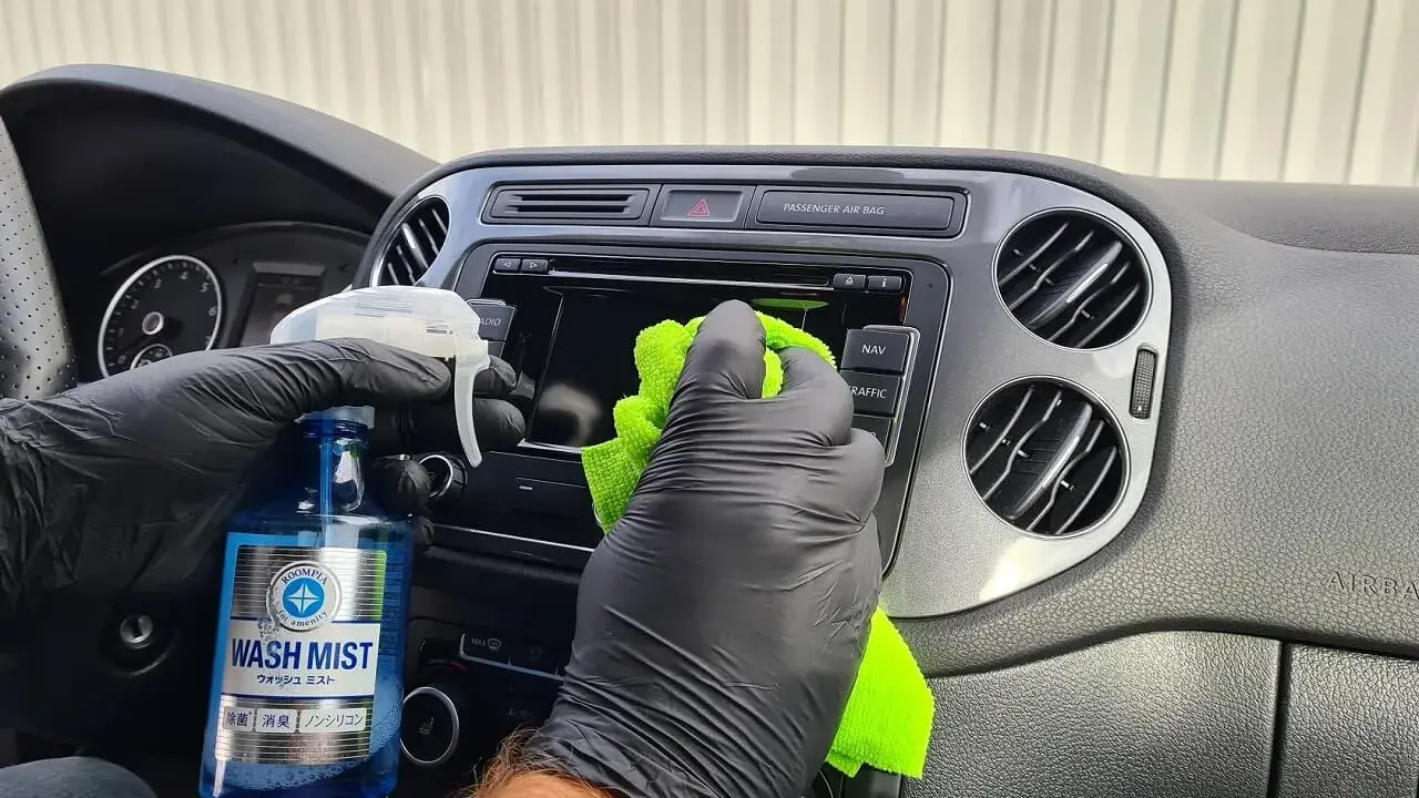 Ręce w czarnych rękawiczkach czyszczą deskę rozdzielczą samochodu. Pokazano, jak wypolerować plastik domowym sposobem, używając płynu "Wash Mist".