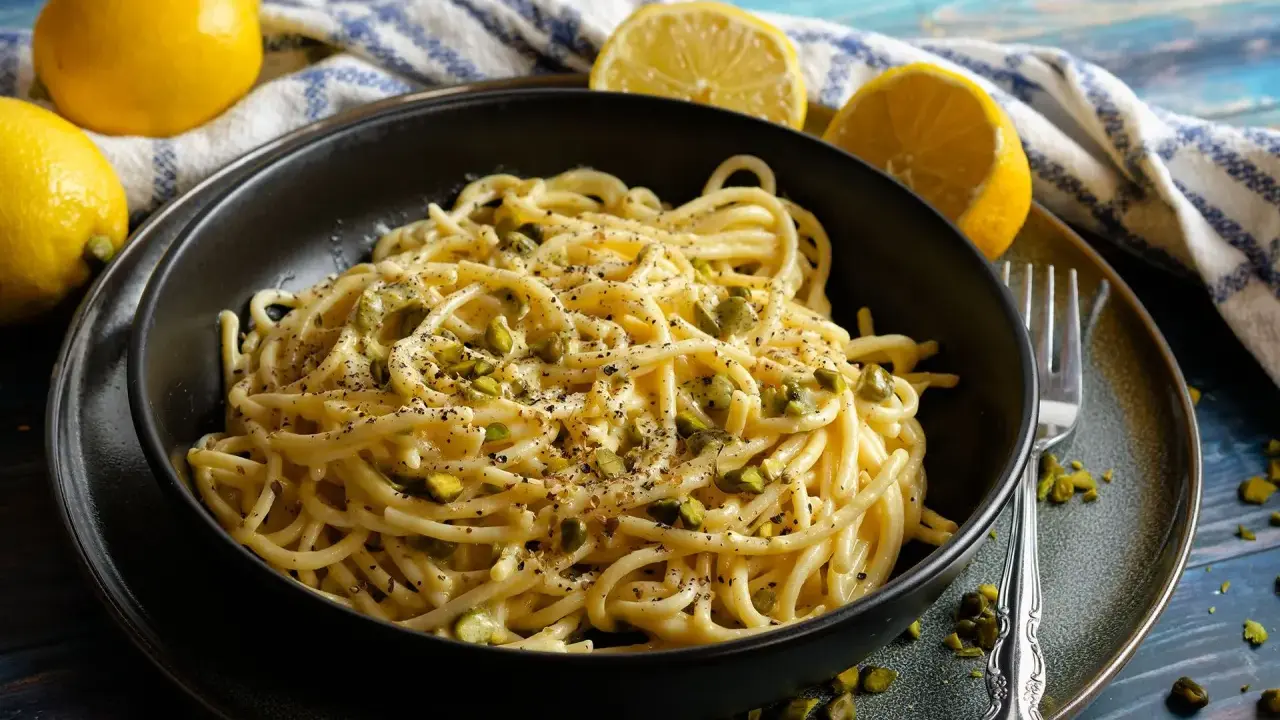 Eine Schüssel mit Zitronen Parmesan Pasta, garniert mit gehackten Pistazien und schwarzem Pfeffer. Frische Zitronen liegen daneben.