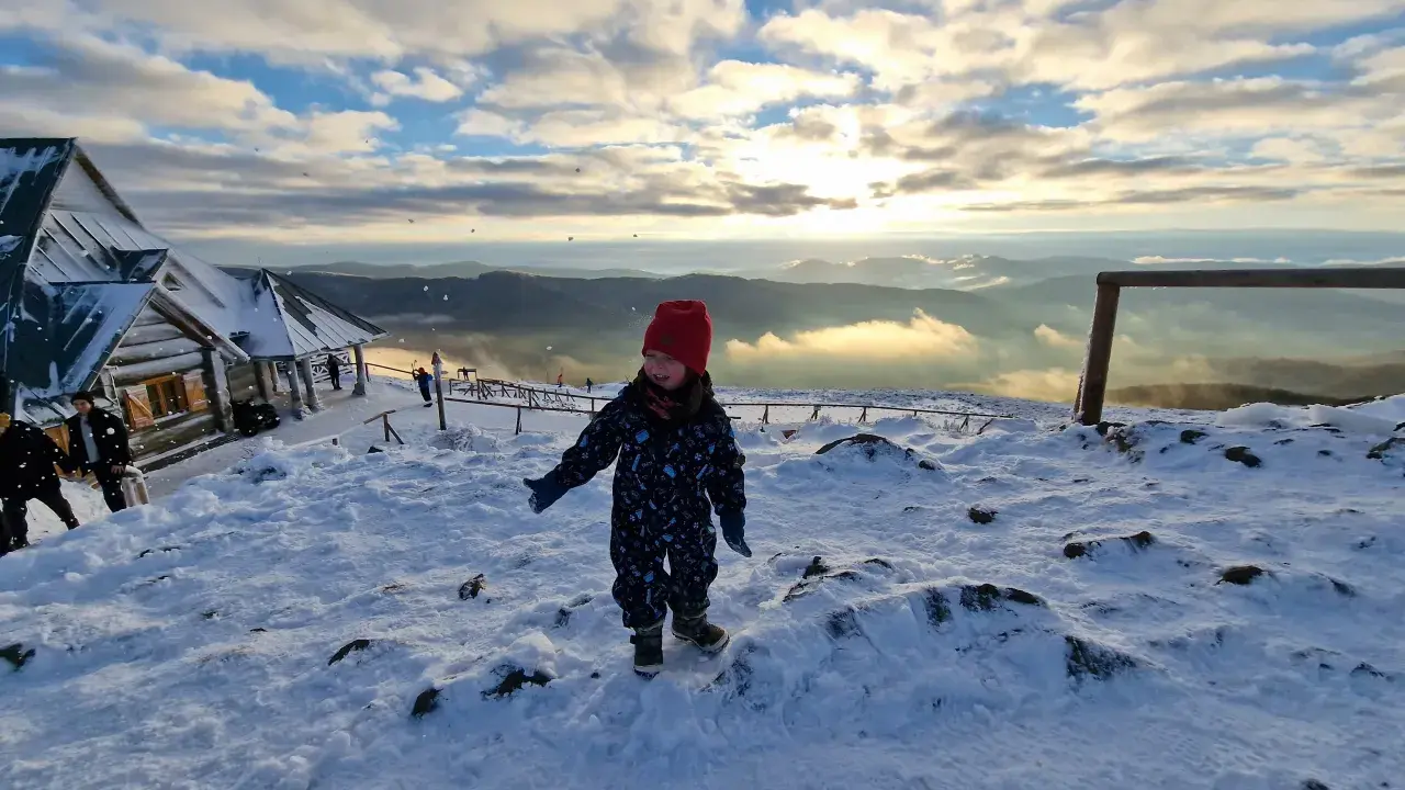 Radosne bieszczady z dziećmi. Mała dziewczynka w czerwonej czapce cieszy się śniegiem na szczycie góry, w tle widać schronisko i piękne zachmurzone niebo.