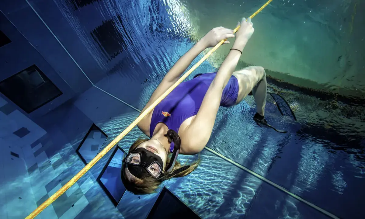 Kobieta w fioletowym stroju kąpielowym i masce nurkuje w basenie, trzymając się liny. To przykład tego, freediving co to jest – sport wymagający kontroli oddechu i siły.
